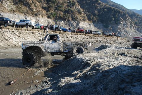 1996 Ford Bronco - A heavily modified pickup truck navigates a muddy off-road course with spectators watching from a nearby embankment.