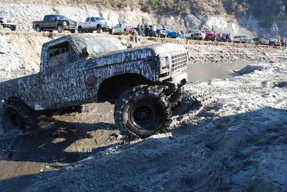Mud-covered 1979 Ford F-Series pickup truck stuck in mud, other trucks and people on embankment behind