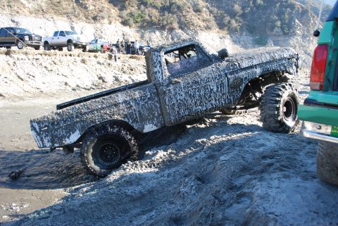 1996 Ford Bronco - This heavily mud-caked pickup truck appears to be stuck in a sandy, wet terrain, with other vehicles nearby.