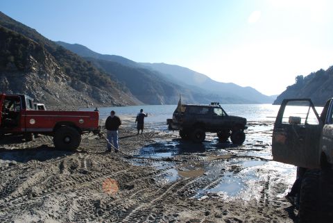 1996 Ford Bronco - Off-road vehicles are parked by a lake with mountains in the background, one truck is being pulled by a rope.