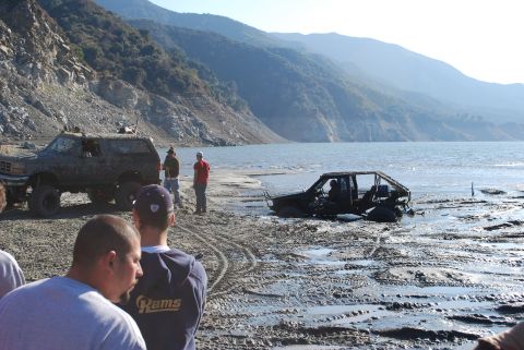 1996 Ford Bronco - Two off-road vehicles are stuck in the mud near a body of water, with people standing around them.