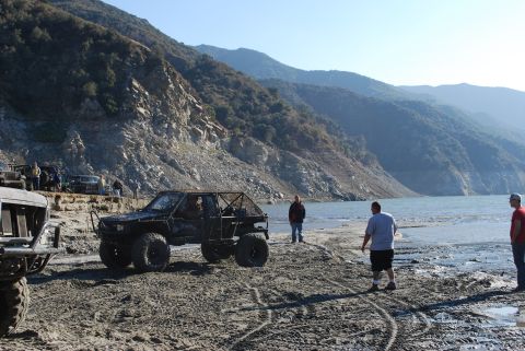 1996 Ford Bronco - Several off-road vehicles are parked on a dirt area near a body of water with mountains in the background.