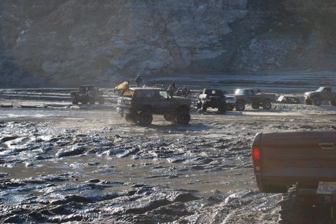 1996 Ford Bronco - Several off-road vehicles navigate a muddy terrain under a rocky cliff face.