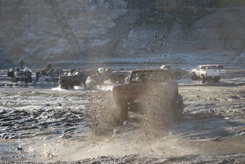 1996 Ford Bronco - Multiple off-road vehicles navigate a muddy, rocky landscape, kicking up large sprays of dirt and water.