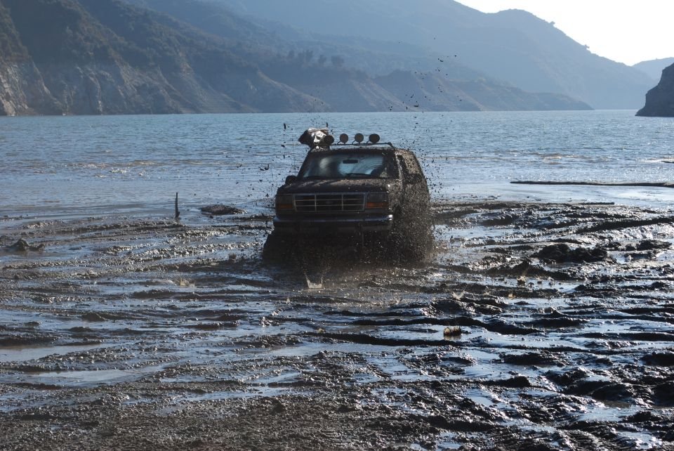 Brown Ford Bronco driving through mud and water, throwing up spray. Roof-mounted lights are visible