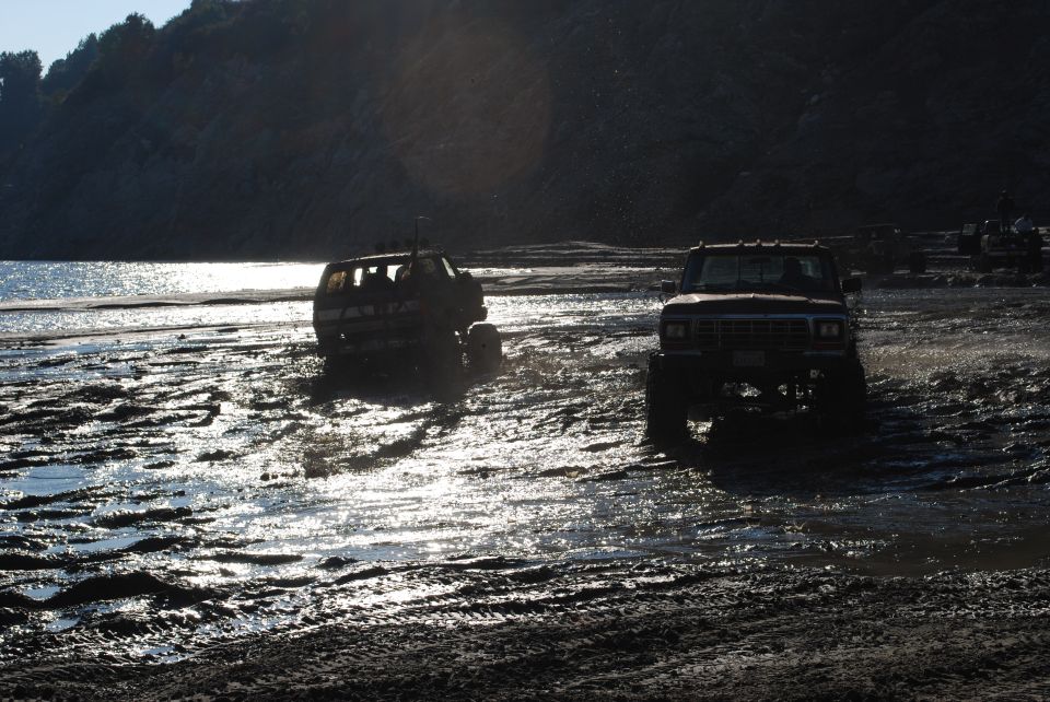 Two lifted trucks driving through a muddy, wet area with a body of water and hills in the background
