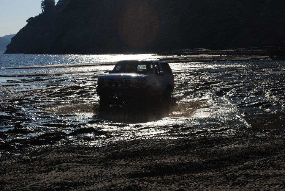 Dark SUV driving through mud and water near a hill and ocean