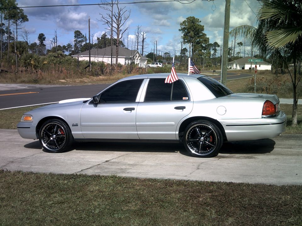 Silver Ford Crown Victoria sedan side view with black aftermarket wheels, tinted windows, and two American flags