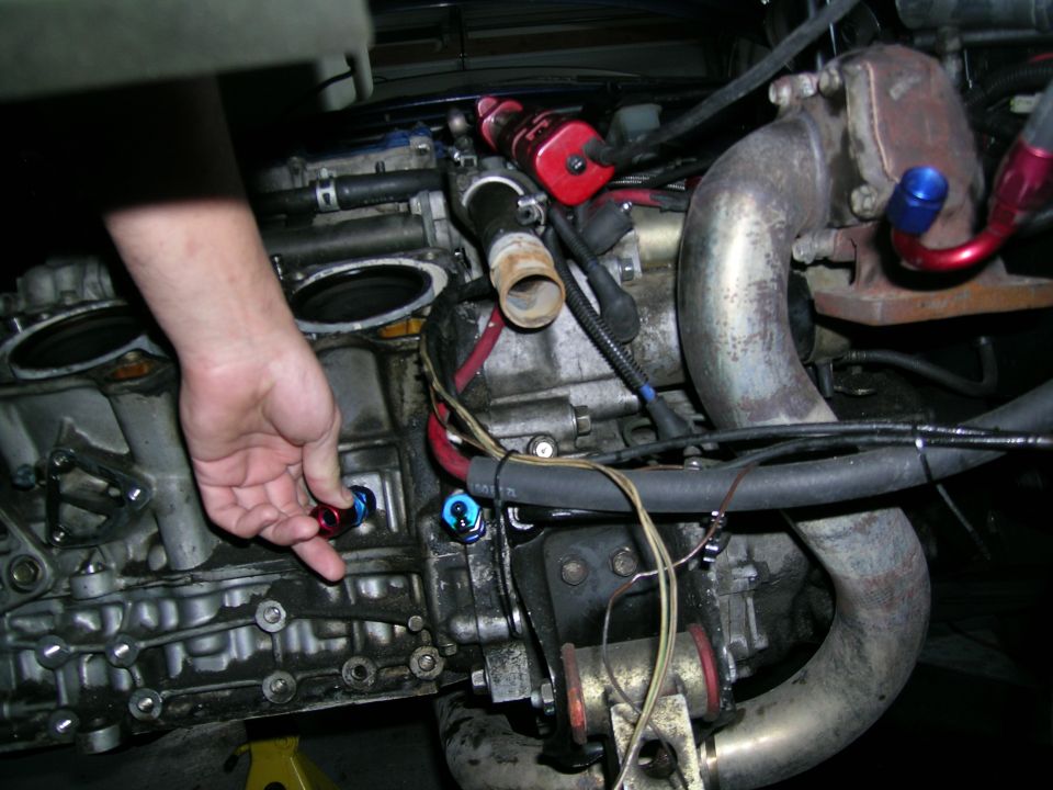 Engine block with hand touching a red and blue fitting, turbocharger, and plumbing