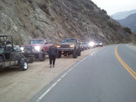 1979 Ford Bronco - Several lifted trucks and a go-kart on a trailer are parked on the side of a mountain road.