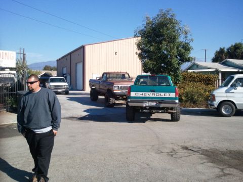 1979 Ford Bronco - Rear view of a lifted teal Chevrolet pickup truck parked next to a brown Ford pickup and a white van.