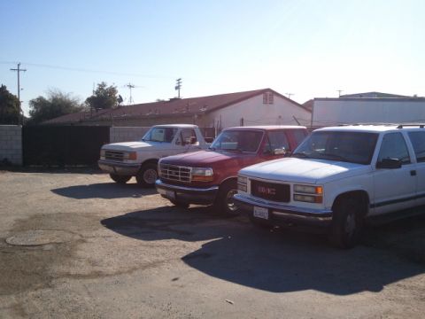 1979 Ford Bronco - Three trucks, a white Ford F-Series, a red Ford Bronco, and a white GMC Suburban, are parked in a lot under a clear blue sky.
