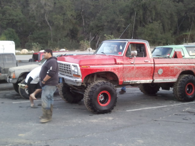 Red lifted pickup truck with big tires parked next to another truck. Someone is looking under the hood of the other truck