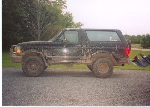 1996 Ford Bronco - Driver-side profile of a black full-size Ford Bronco heavily splattered with dried mud, sitting on a gravel driveway with green lawn and tr…