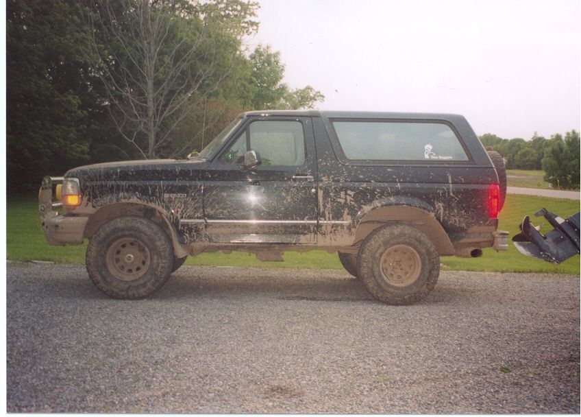 Mud-covered black full-size Ford Bronco in side profile on gravel driveway with brush guard