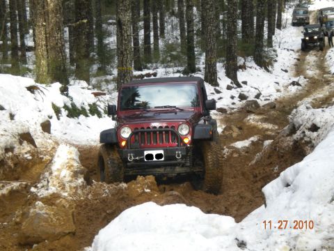 2005 Jeep Wrangler - A red 2005 Jeep Wrangler tackles a challenging muddy and snowy off-road course, with other vehicles visible in the background.