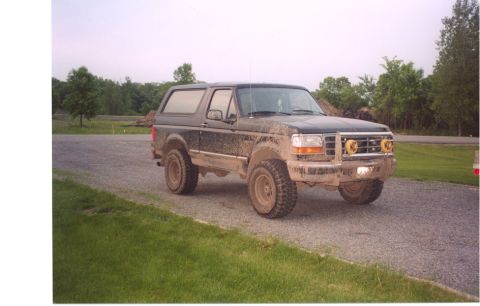 1996 Ford Bronco - Three-quarter front view of a mud-caked black full-size Ford Bronco parked on a gravel drive beside a grass shoulder, wearing a tube brush…