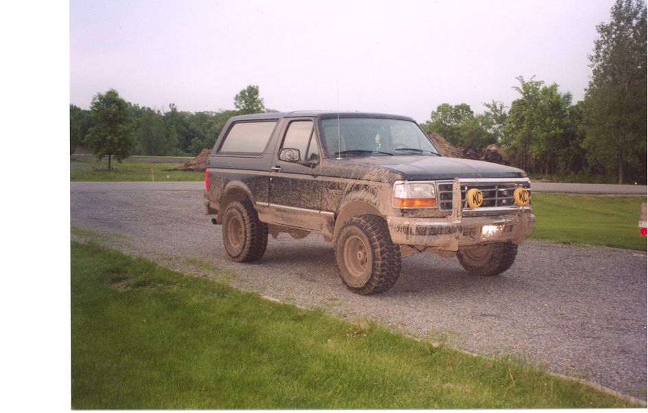 Muddy black Ford Bronco 3/4 front on gravel drive with brush guard and yellow KC lights