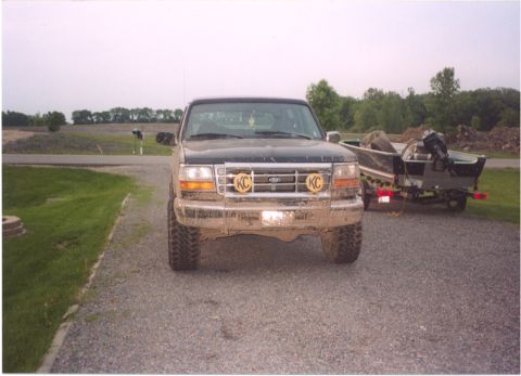1996 Ford Bronco - lifted Ford truck wearing yellow KC driving lights, hitched to a boat trailer on a gravel parking area.