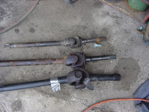 1992 Ford Bronco - Three front axle shafts laid side by side on a shop floor for comparison, each with a U-joint and splined stub of visibly different diamete…