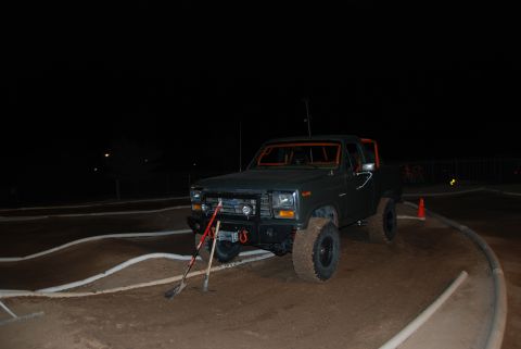 1982 Ford Bronco - This dark green SUV, likely from the early 1980s, features an orange roll cage and is parked on a dirt track at night.