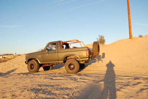 1982 Ford Bronco - Olive green Ford Bronco with an orange roll cage and spare tire on the rear is seen from a 3/4 front angle on a dirt hill.