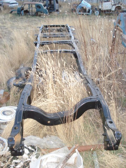 1977 Ford F-150 - A bare truck frame sits in a field of dry grass, surrounded by other discarded vehicle parts and rusted car bodies.