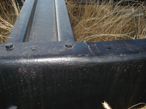 1977 Ford F-150 - Detail of a truck frame, showing bolted sections and surrounding dry vegetation. A website logo is present.