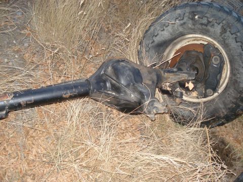1977 Ford F-150 - A detached rear axle assembly and tire lie on the ground amidst dry grass and brush.