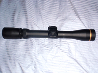 1991 Ford Bronco - A black rifle scope with a gold ring and visible branding is shown against a white, textured background.