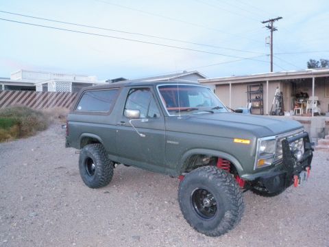 1982 Ford Bronco - Olive green Ford Bronco with aftermarket bumper and lifted suspension sits on a gravel lot.