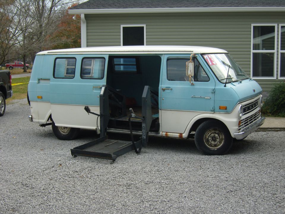 Two-tone blue and white van with a deployed wheelchair lift, parked on a gravel driveway