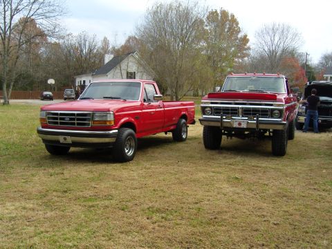 2007 Ford Mustang - Two red pickup trucks are parked on a dry, grassy lawn in front of a house.