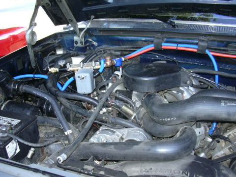 1991 Ford Bronco - Close-up into the engine bay of a dark blue Ford truck with the hood raised, showing a carbureted V8 wearing a black round air cleaner, thi…