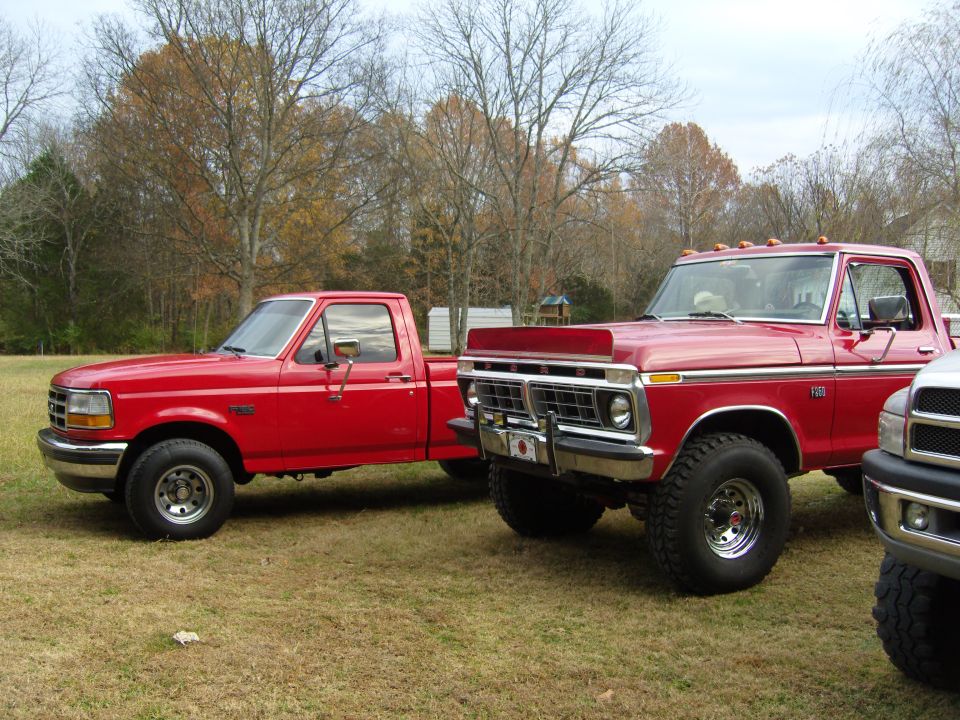 Two red pickup trucks parked on grass. One is a newer model Ford F-150, the other is an older model Ford F-250