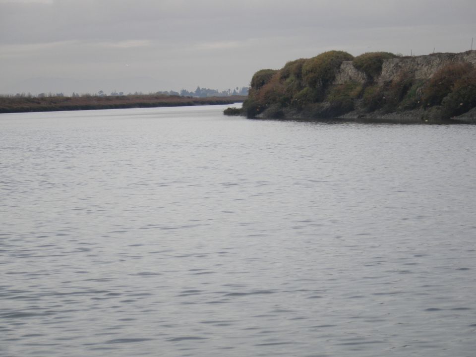 Waterway with a rocky, vegetated embankment under an overcast sky. Supermotors.net logo in lower right