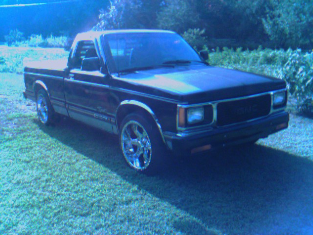 Black GMC compact pickup on chrome wheels, front three-quarter view, parked on grass