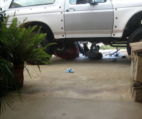 1996 Ford Bronco - Two individuals are working on the underside of a white Ford Bronco, with tools laid out nearby.