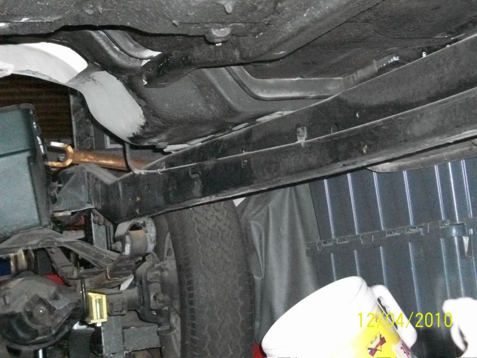Underside view of a vehicle's frame, suspension, and fuel tank. A tire is visible