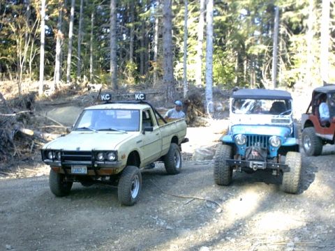 1995 Ford Bronco - Two off-road vehicles, a pickup truck and a Jeep, are parked on a dirt road in a wooded area.