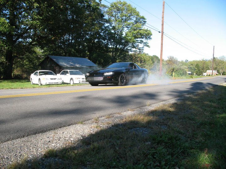 Black 2002 Chevrolet Camaro doing a burnout on a road, two white cars parked nearby