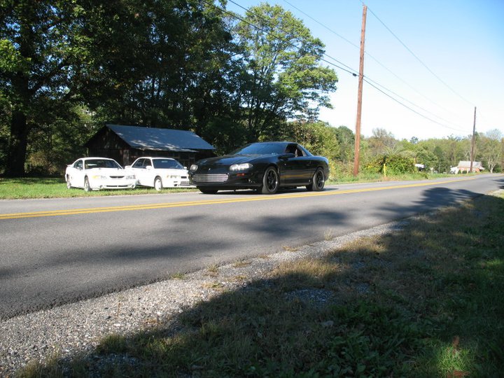Black 2002 Chevrolet Camaro driving on a road, with two white cars parked on the side