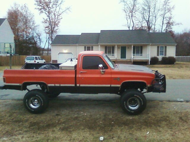 Lifted orange and black square-body pickup truck, side view, parked in front of a house