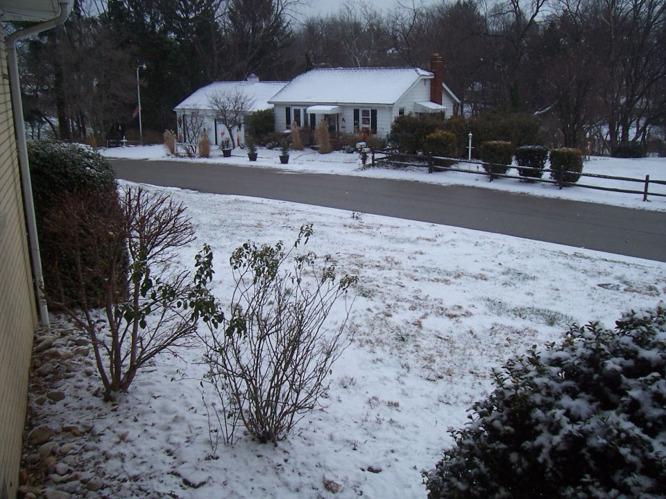 Snow-covered residential street with a white house and bare trees