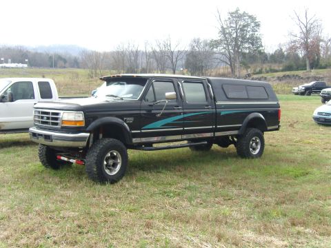 2007 Ford Mustang - Lifted black 1992 Ford F-350 pickup truck with a camper shell and teal graphics is parked in a field.