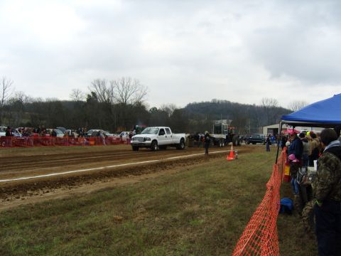 2007 Ford Mustang - White pickup truck competes in a pull event, dragging a sled on a dirt track with spectators watching.
