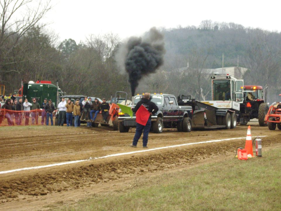 Dark blue pickup truck with dual rear wheels pulling a sled, emitting black smoke