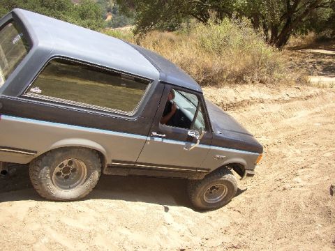1989 Ford Bronco - This 1989 Ford Bronco is navigating a dusty, off-road path with dry brush and trees in the background.
