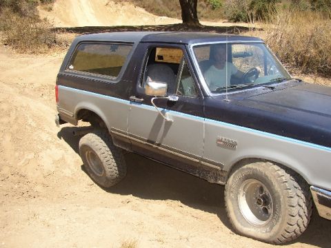 1989 Ford Bronco - A two-tone Ford Bronco is driven on a dirt path, with a driver visible inside.