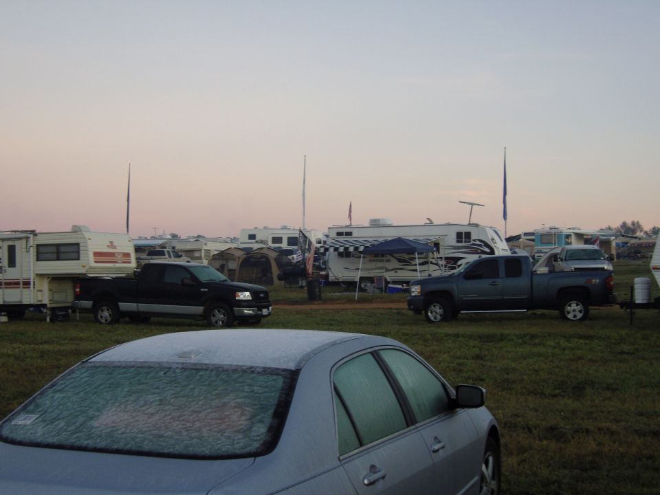 Silver sedan rear view with frost on the windshield and roof, parked in a grassy field with RVs and trucks in the background